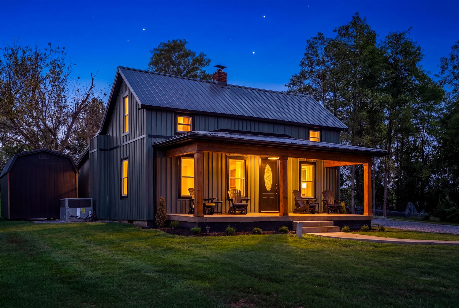 The dark green board-and-batten farmhouse at dusk, windows lit and chairs on the covered porch.