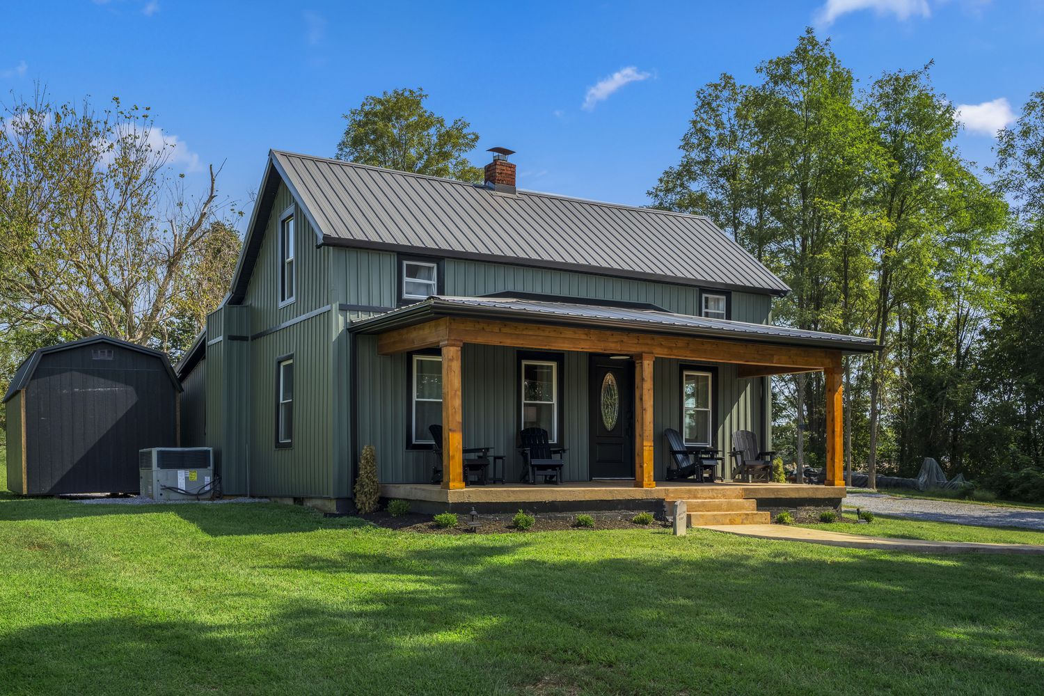 Front of the farmhouse in daylight: dark green board-and-batten, standing-seam metal roof, cedar porch posts.