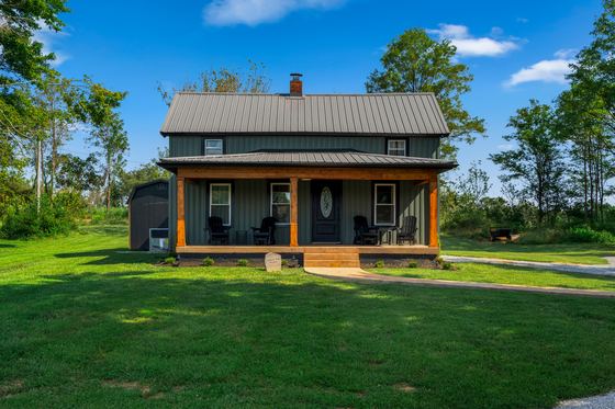 The farmhouse from the side, showing the full length of the porch and the metal roof.
